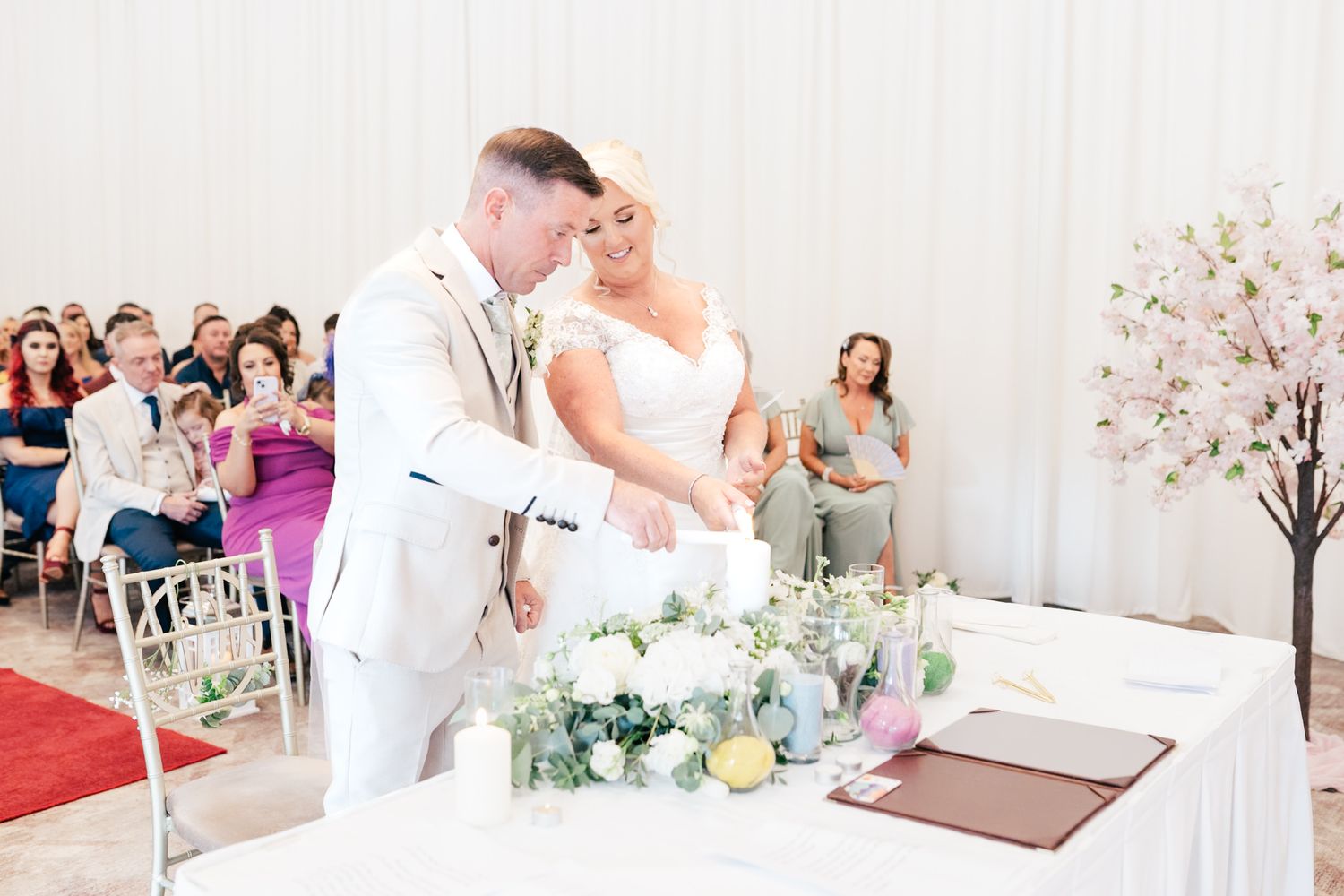 A couple signs their marriage certificate at a wedding ceremony with white decor and cherry blossoms in the background.