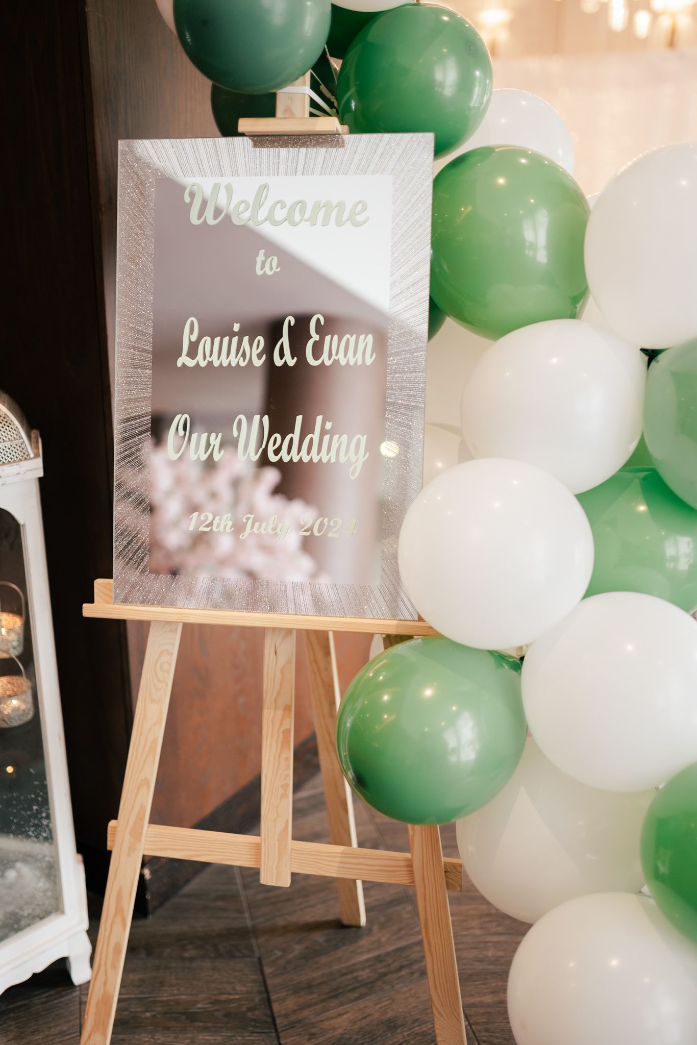 Wedding welcome sign on wooden easel with green and white balloon arch backdrop displaying Louise and Evan's names.