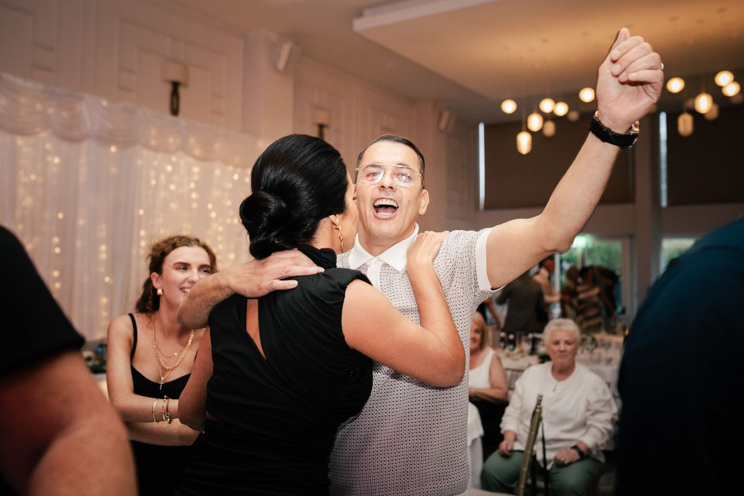 Wedding guests energetically dancing and celebrating at an indoor reception with string lights and white decor.