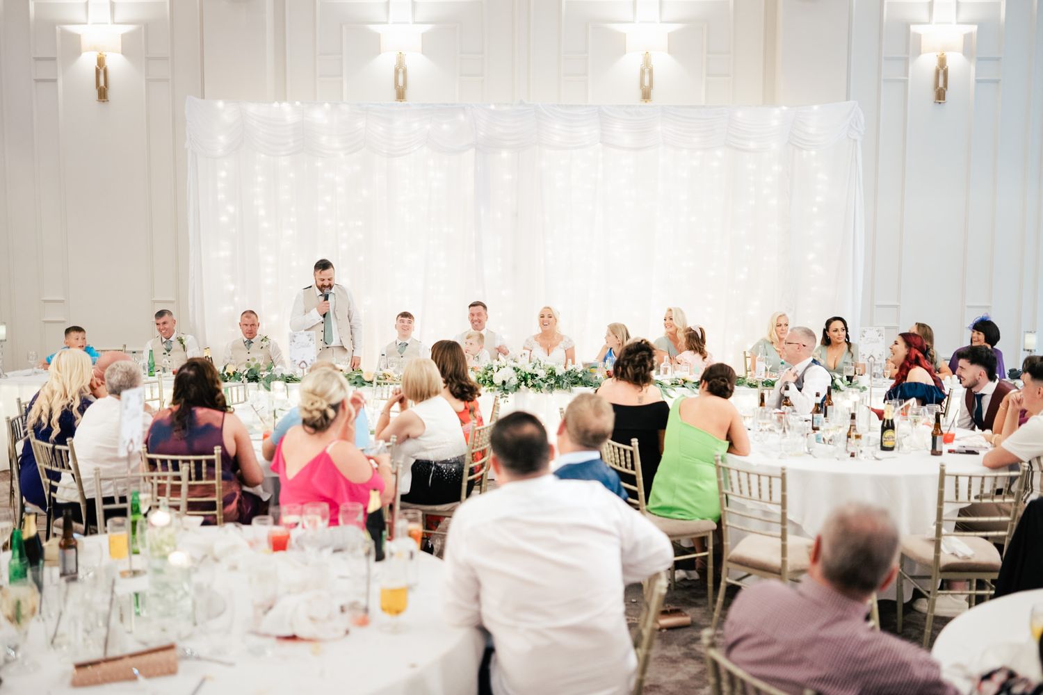 Wedding reception guests seated at elegant tables with white decor enjoy speeches during a celebration in a bright ballroom.