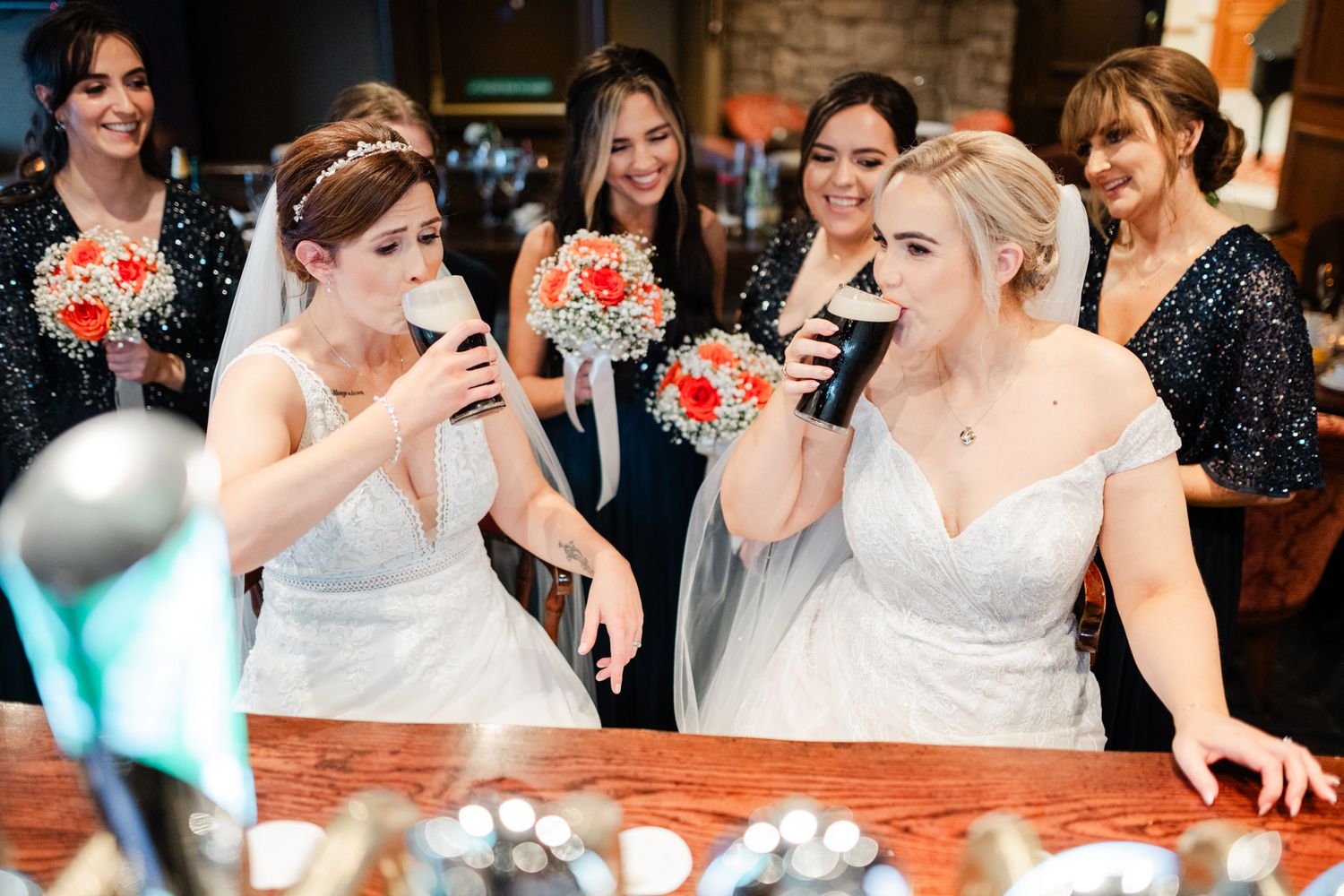 Two people in white wedding dresses share drinks together while celebrating with bridesmaids in dark dresses at a reception.