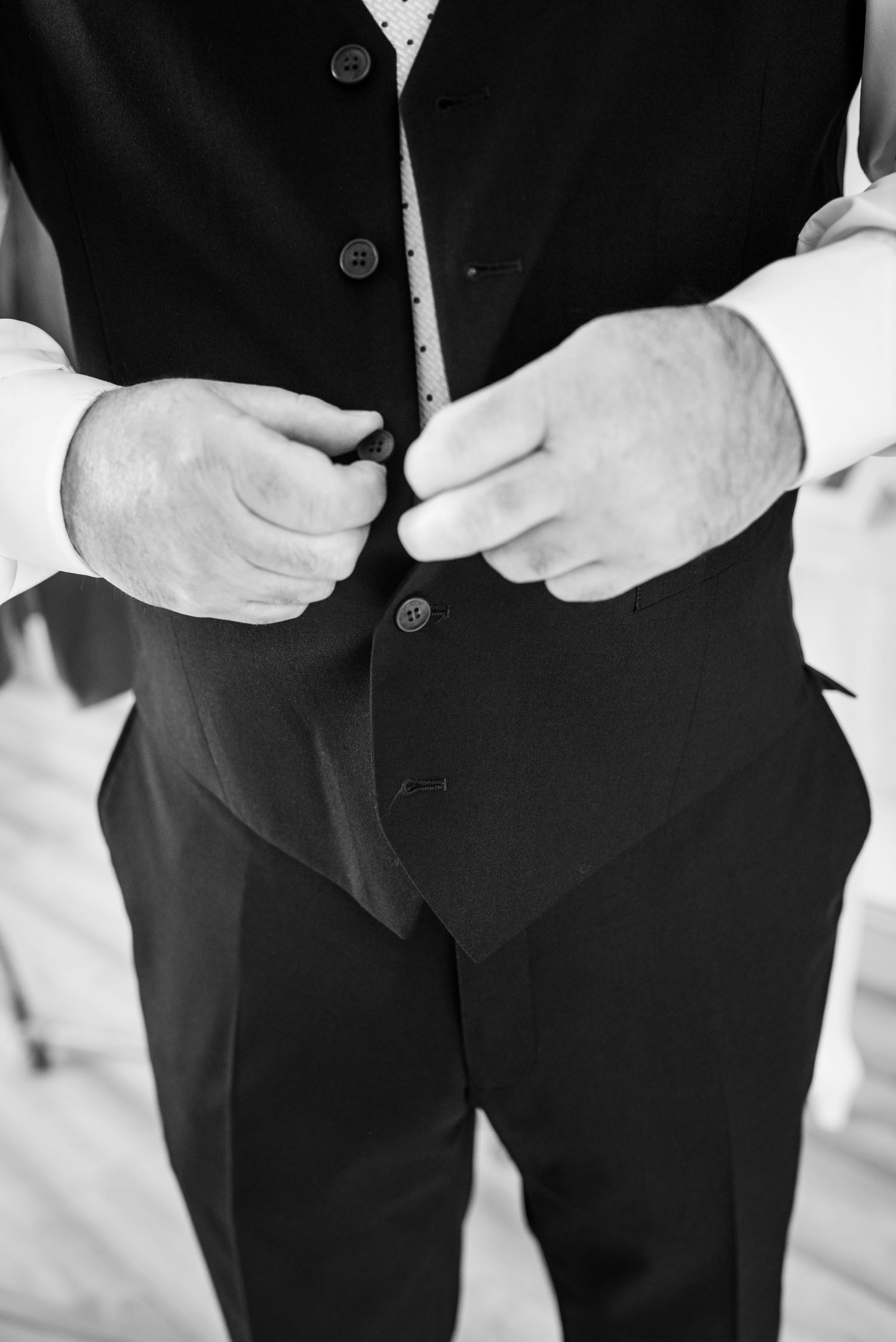 Black and white close-up of hands buttoning up a formal black suit vest during dressing preparation.