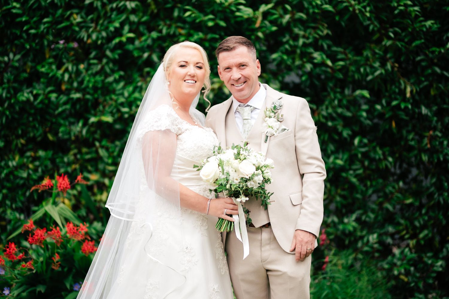 A bride and groom pose together outdoors against lush greenery, with the bride holding a white floral bouquet.