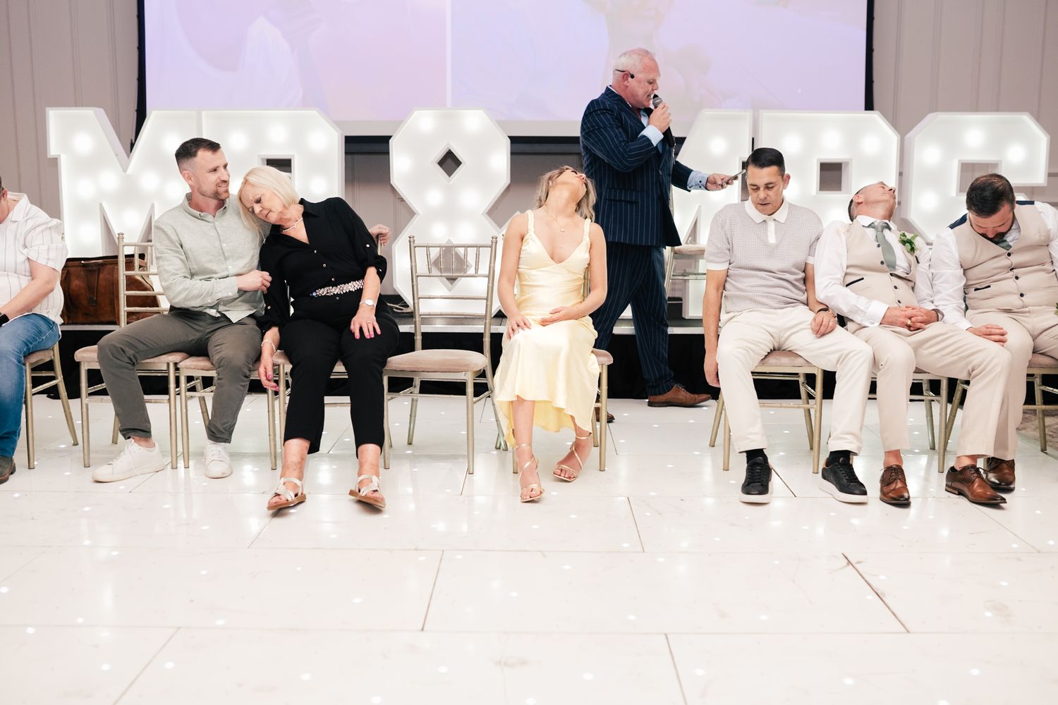 People wearing white clothing sit on chairs along a white wall during an indoor gathering or event.