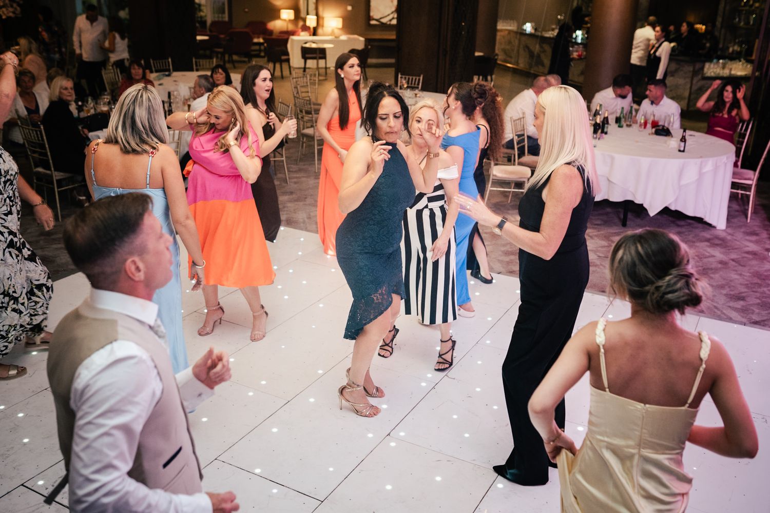 Energetic party guests dance and celebrate on an illuminated white dance floor at an evening reception.