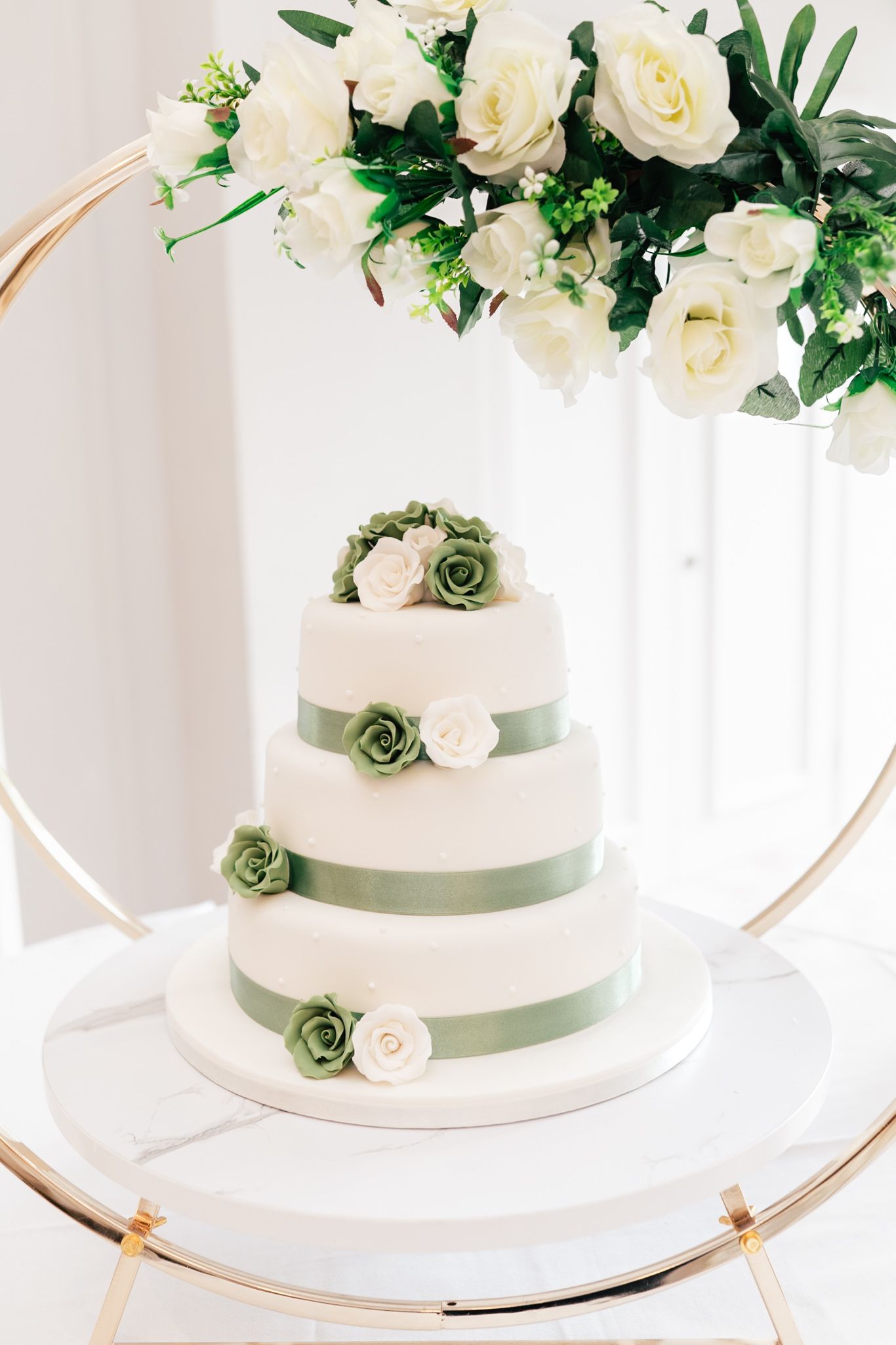 White three-tier wedding cake decorated with cream roses and sage green ribbons on a gold circular stand against light backdrop.