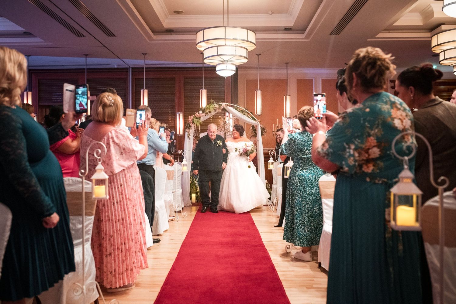 Wedding guests in formal attire line a red carpet aisle at an indoor reception as lanterns illuminate the elegant venue.