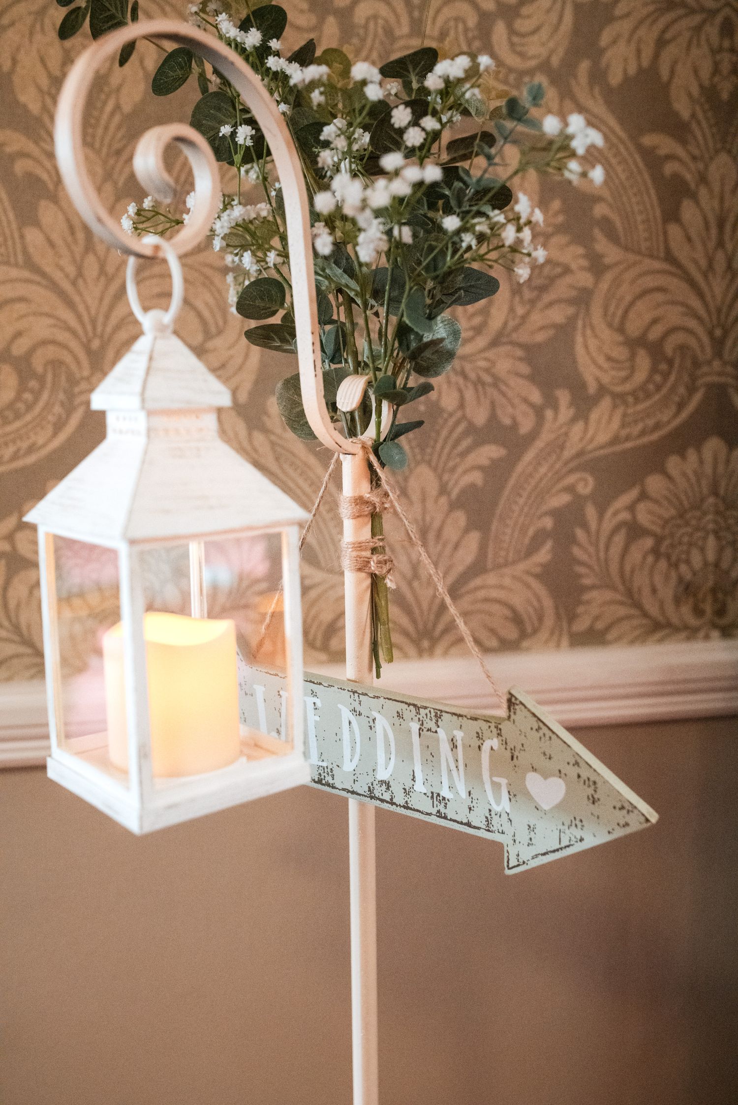 Decorative white lanterns and greenery hang from rustic hooks against a damask patterned wall creating elegant wedding decor.