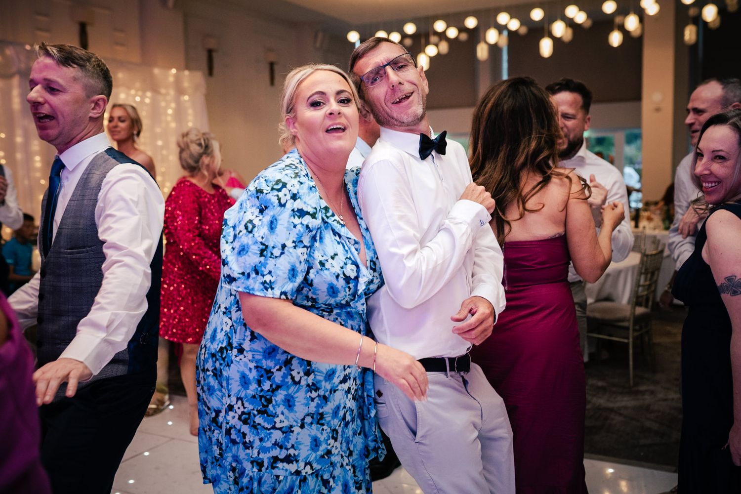 People dancing and celebrating joyfully at a wedding reception with string lights glowing overhead in an indoor venue.