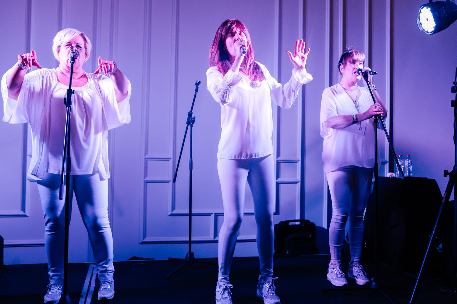 Three performers in white outfits sing on stage under purple lighting during a musical performance.