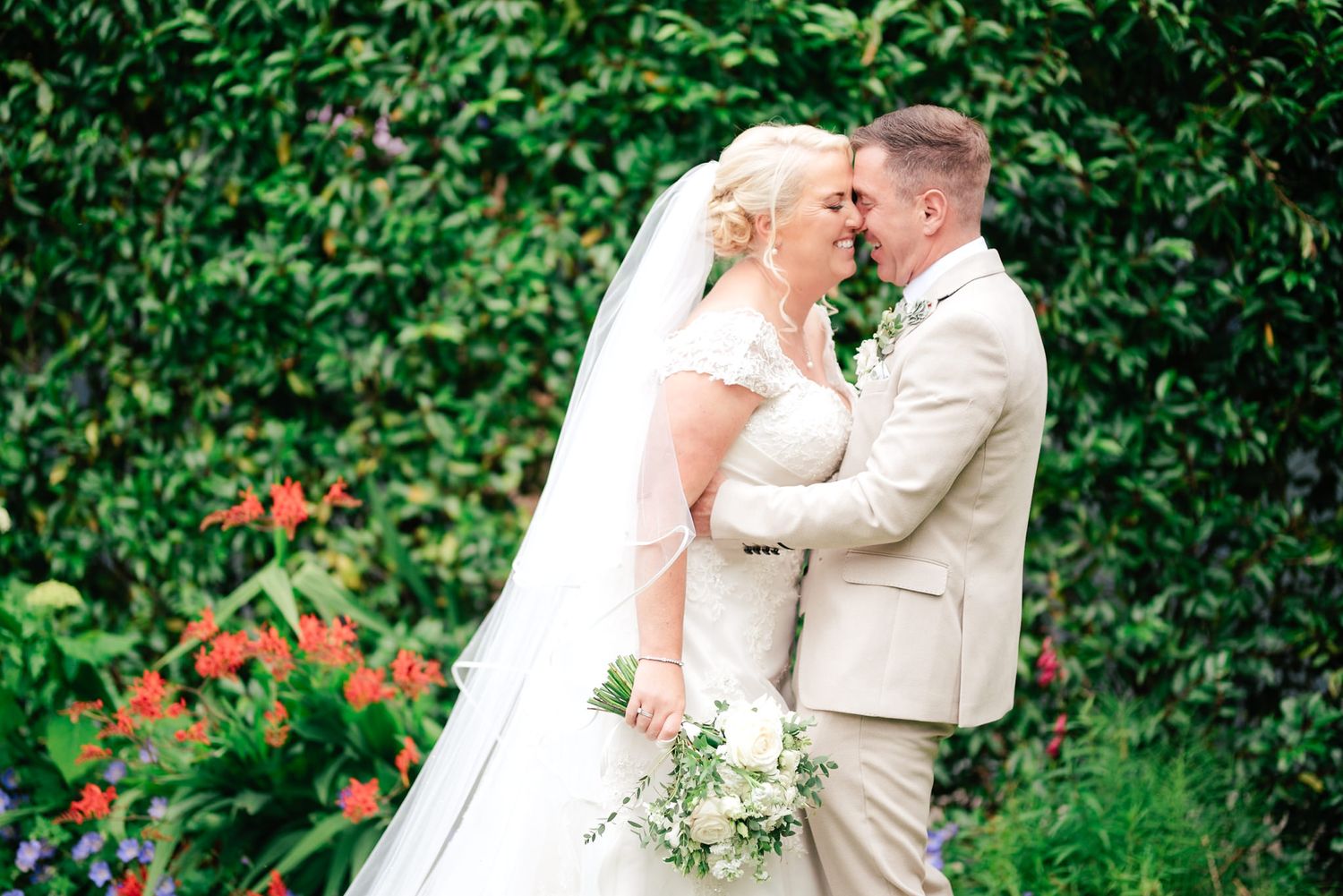 A romantic wedding couple embraces in a garden setting with lush greenery and orange flowers in the background.