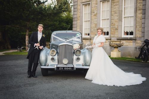 A couple poses with a classic silver Bentley automobile outside a stone building on their wedding day.