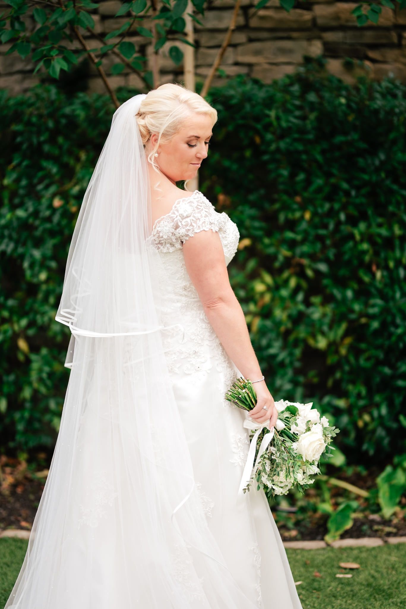 A bride in a white lace gown and long veil holds a bouquet while standing against a leafy garden backdrop.