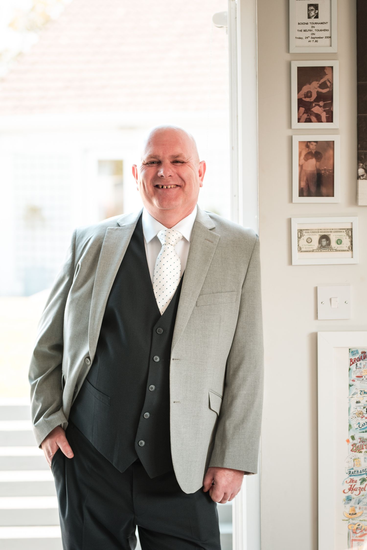 Person in a formal light gray suit jacket and dark vest stands smiling next to a white wall with framed artwork.