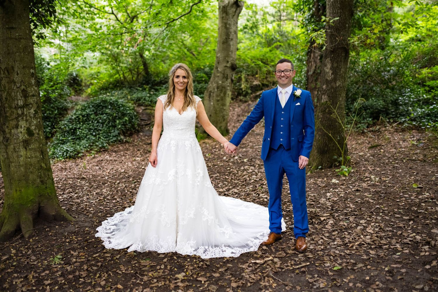Couple in wedding attire holding hands while standing on woodland path covered in fallen leaves.