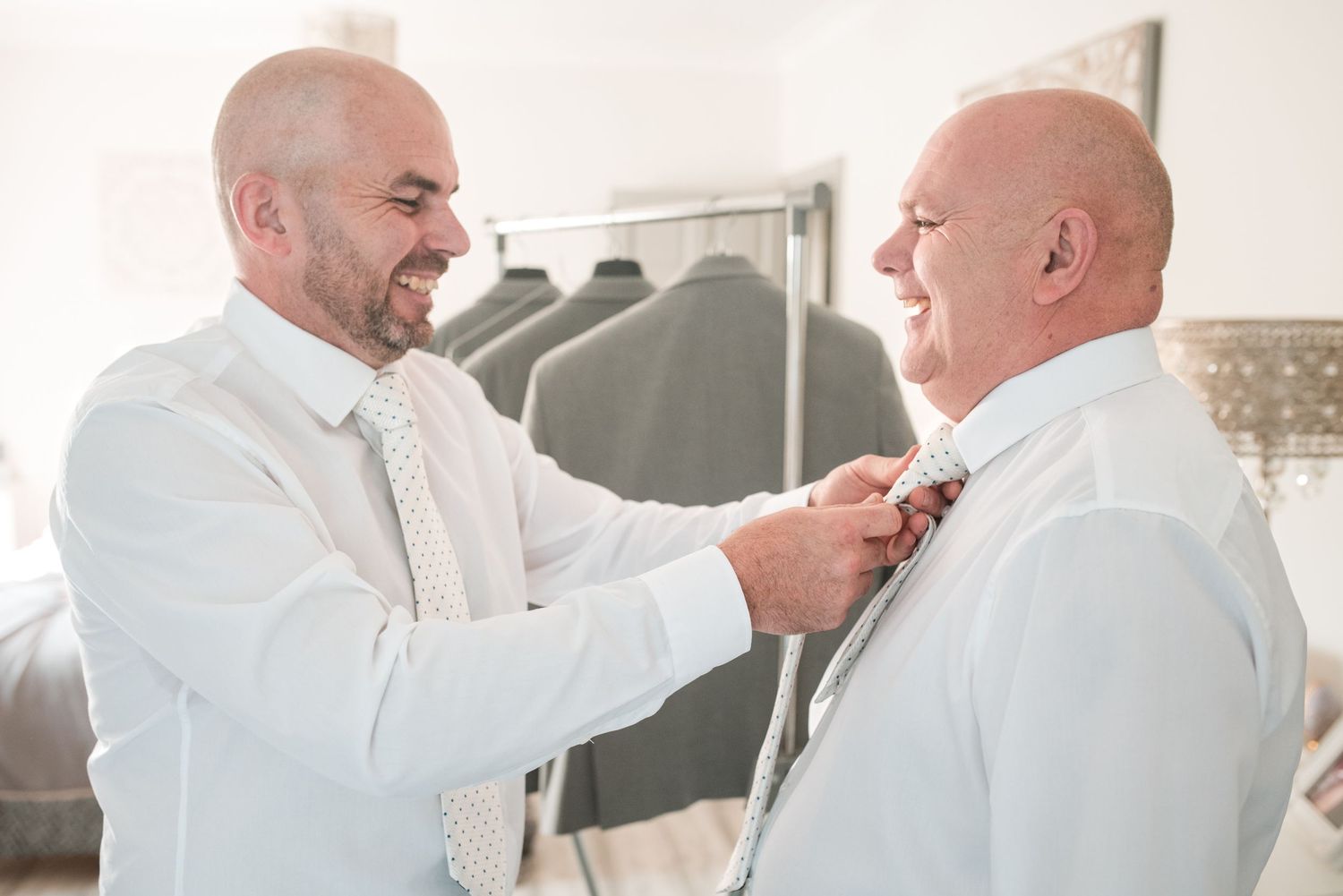Two people in white formal attire share a tender moment as one helps adjust the other's tie indoors.