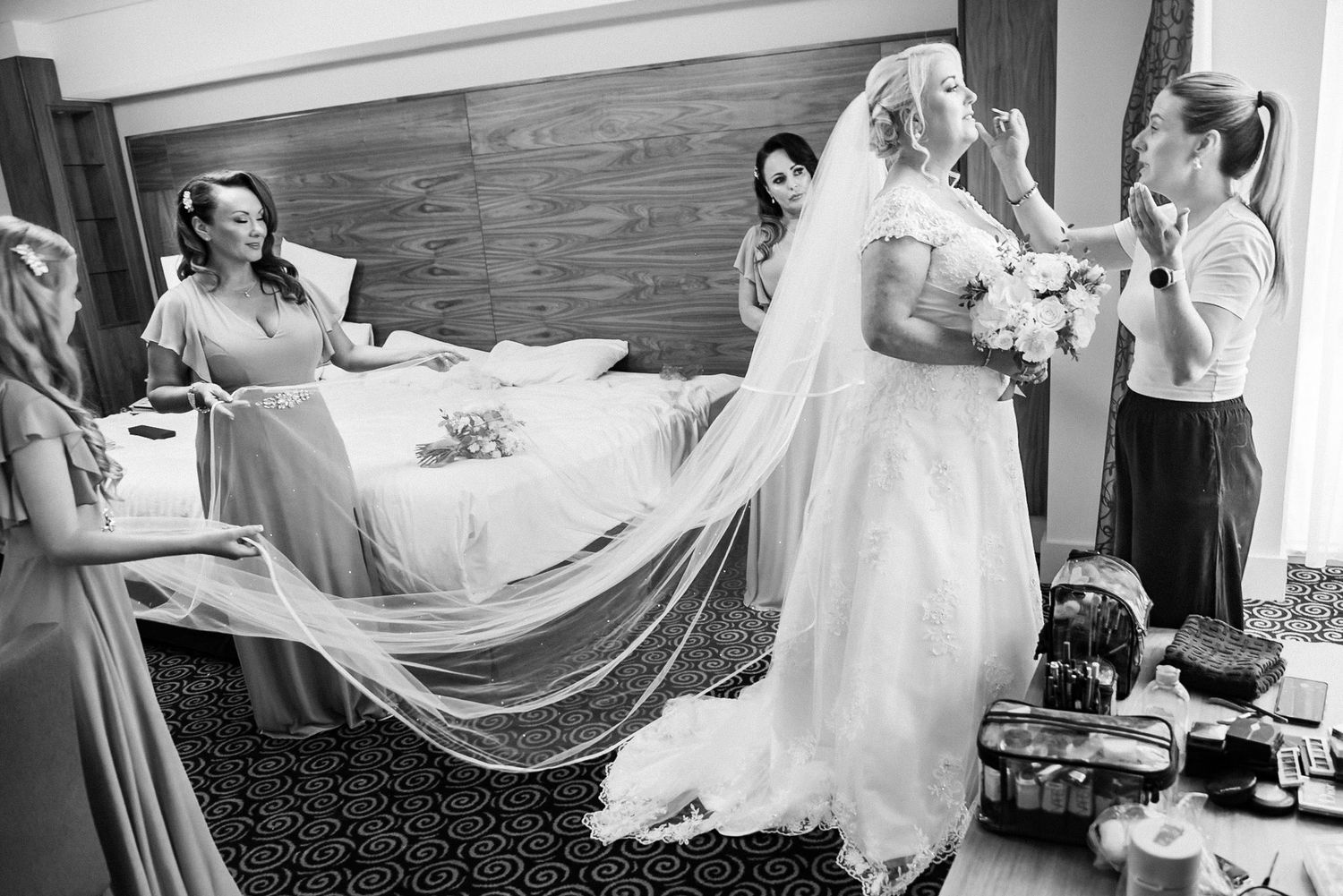 Black and white wedding photo showing a bride in a flowing veil and gown getting ready with helpers in a hotel room.