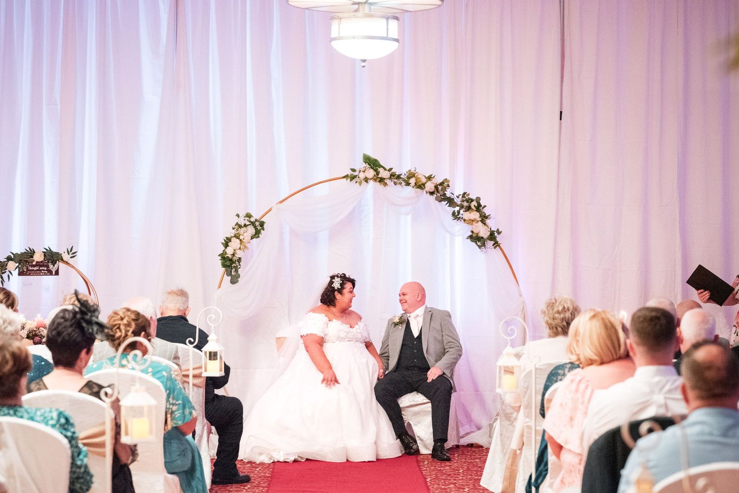 A wedding reception celebration in a white ballroom with hanging chandeliers and guests dancing on the dance floor.