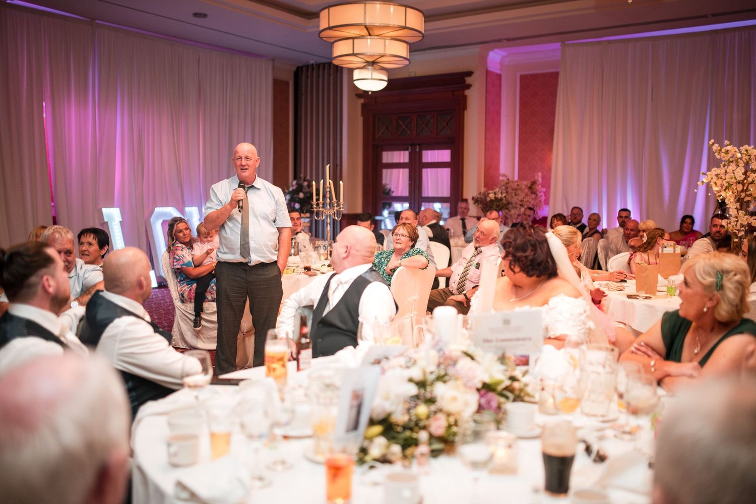 Wedding guests gathered in an elegant reception hall with pink lighting while someone delivers a speech during dinner.
