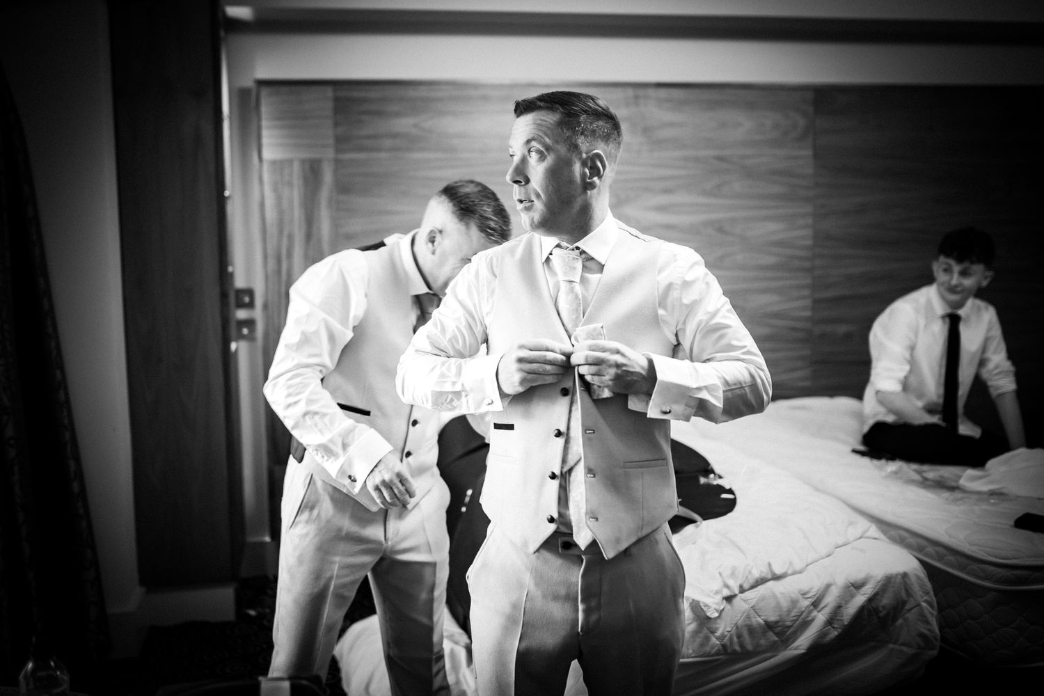 A groom adjusts his vest while getting ready in a black and white hotel room scene.