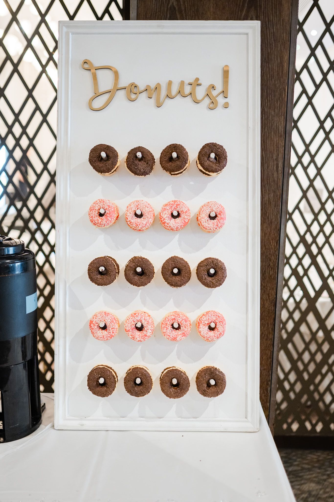 A white pegboard display shows rows of pink and chocolate mini donuts arranged neatly against a lattice backdrop.