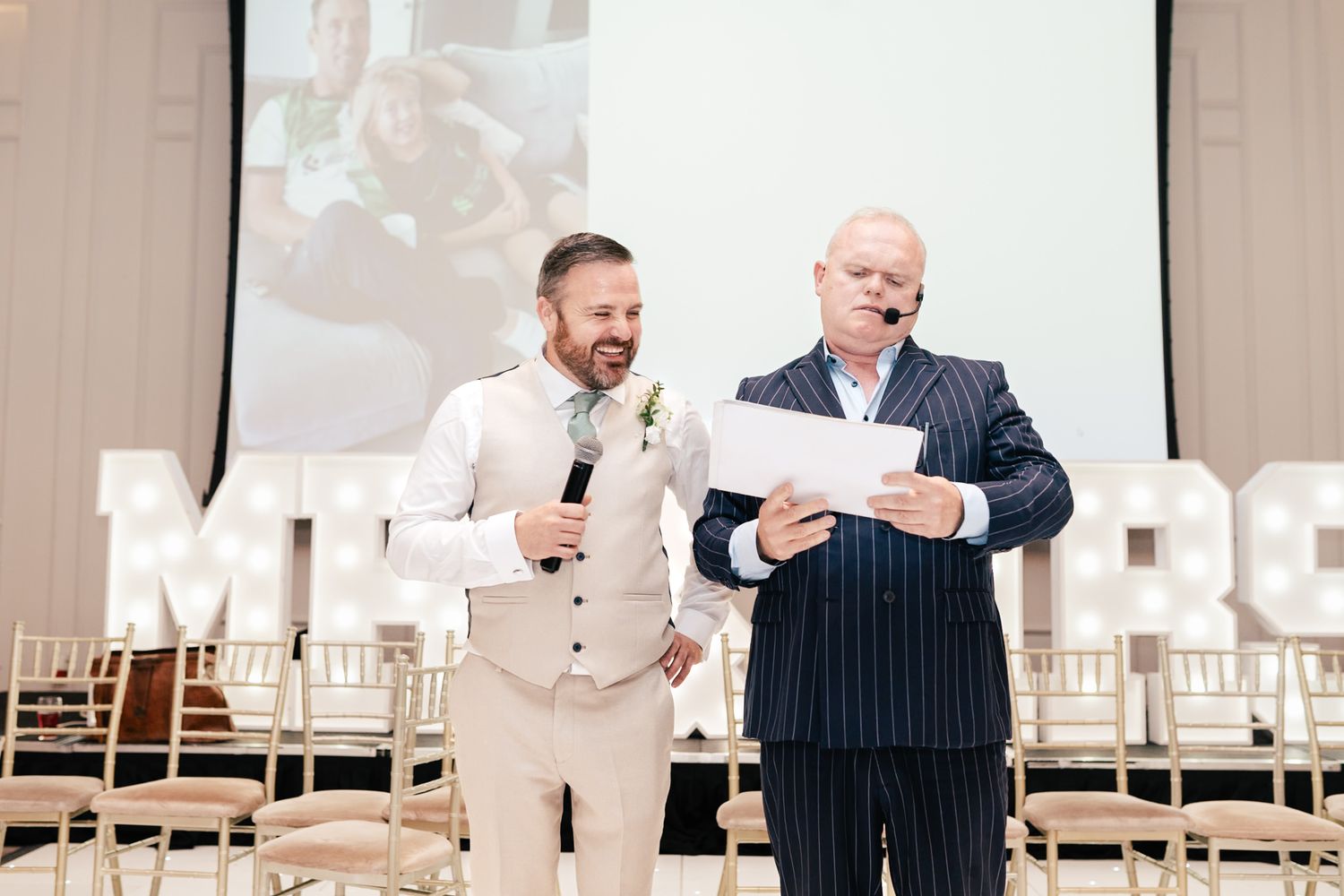 Two people share a laughing moment while standing in an elegant indoor venue with white chairs in the background.