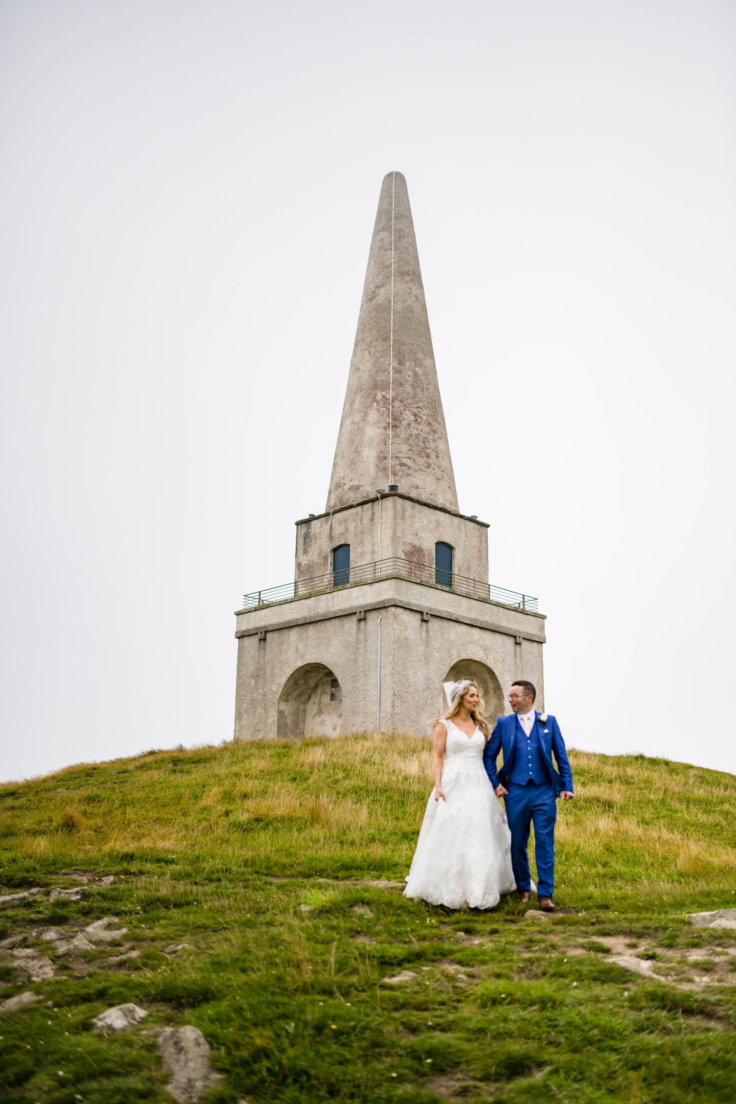A couple taking wedding photos in front of a tall stone obelisk monument on a grassy hilltop.