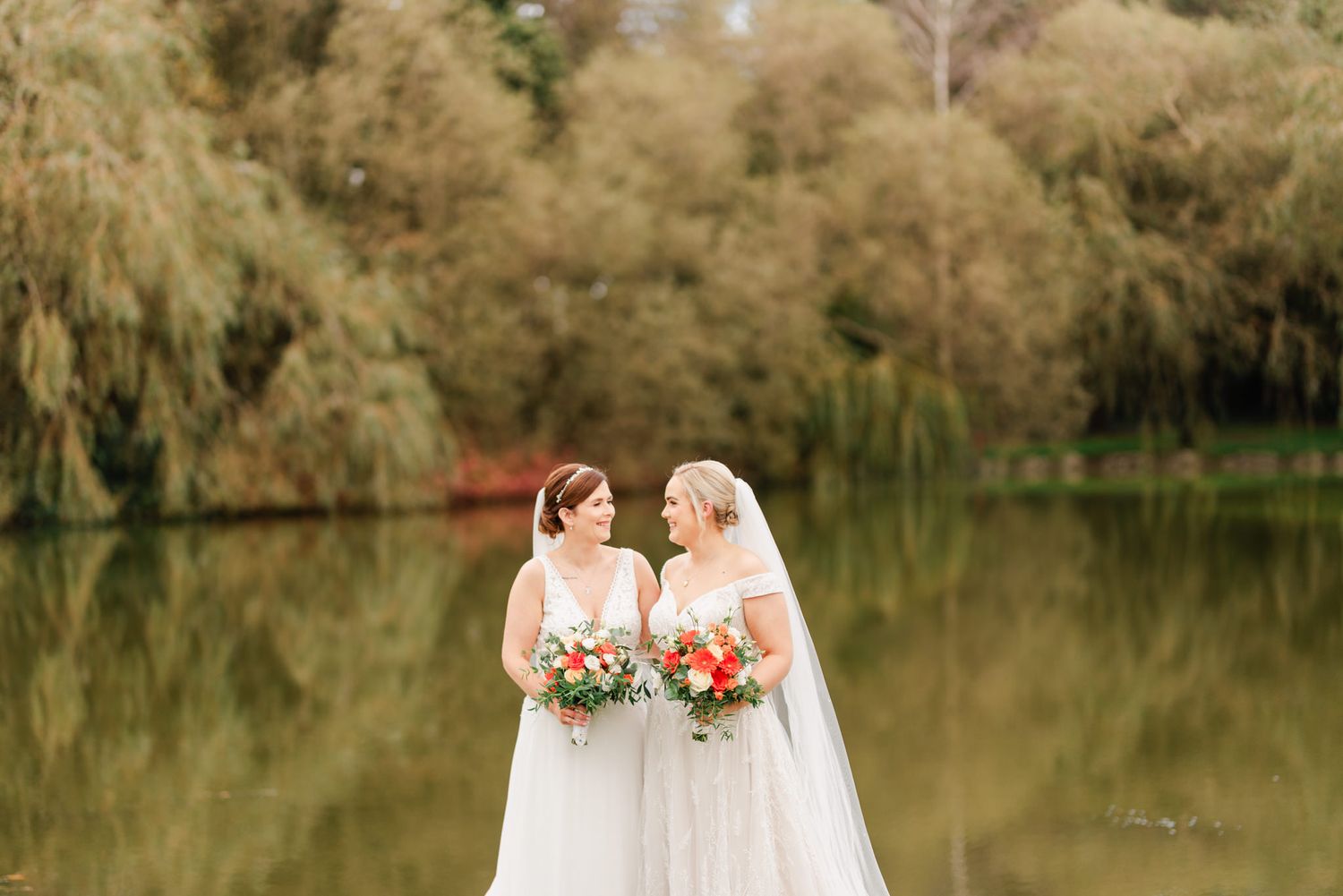Two brides in white wedding dresses hold colorful bouquets while standing by a tranquil lake with weeping willows.