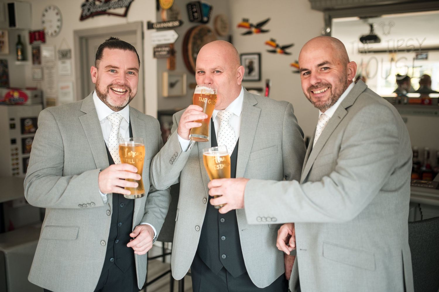 Three groomsmen in matching gray suits toast with beer glasses and smile while celebrating at a wedding venue.