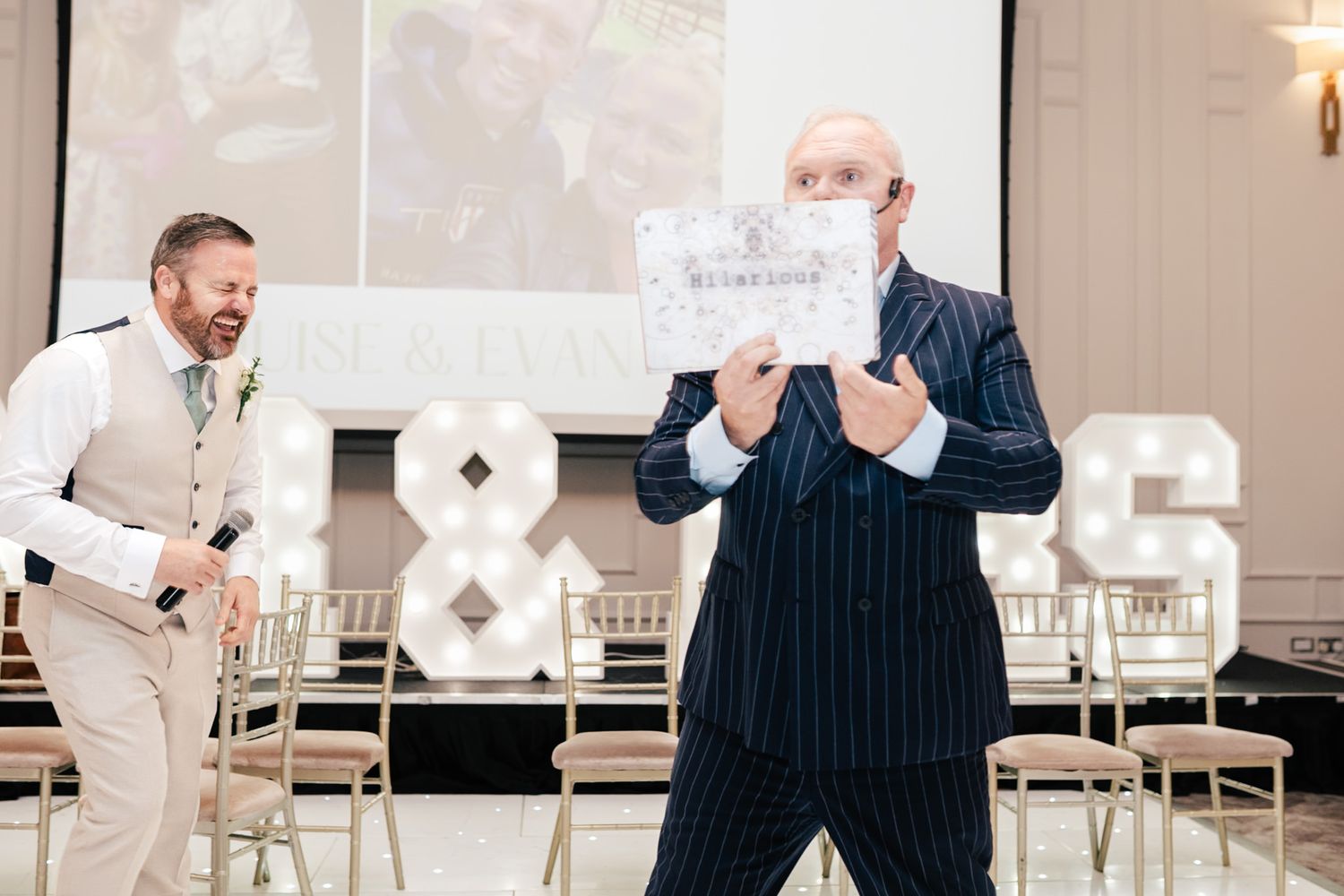 A person in a pinstriped suit holds up a sign while another person in light clothing stands nearby in an event space.