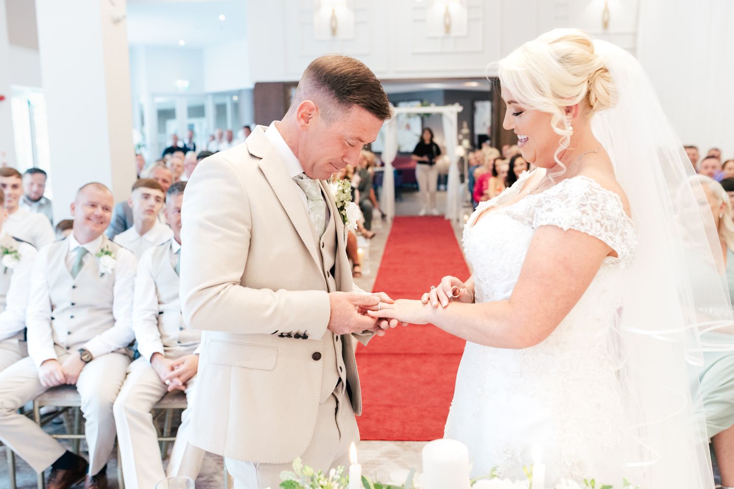 A couple exchanges rings during an outdoor wedding ceremony with guests seated around a red carpet aisle.