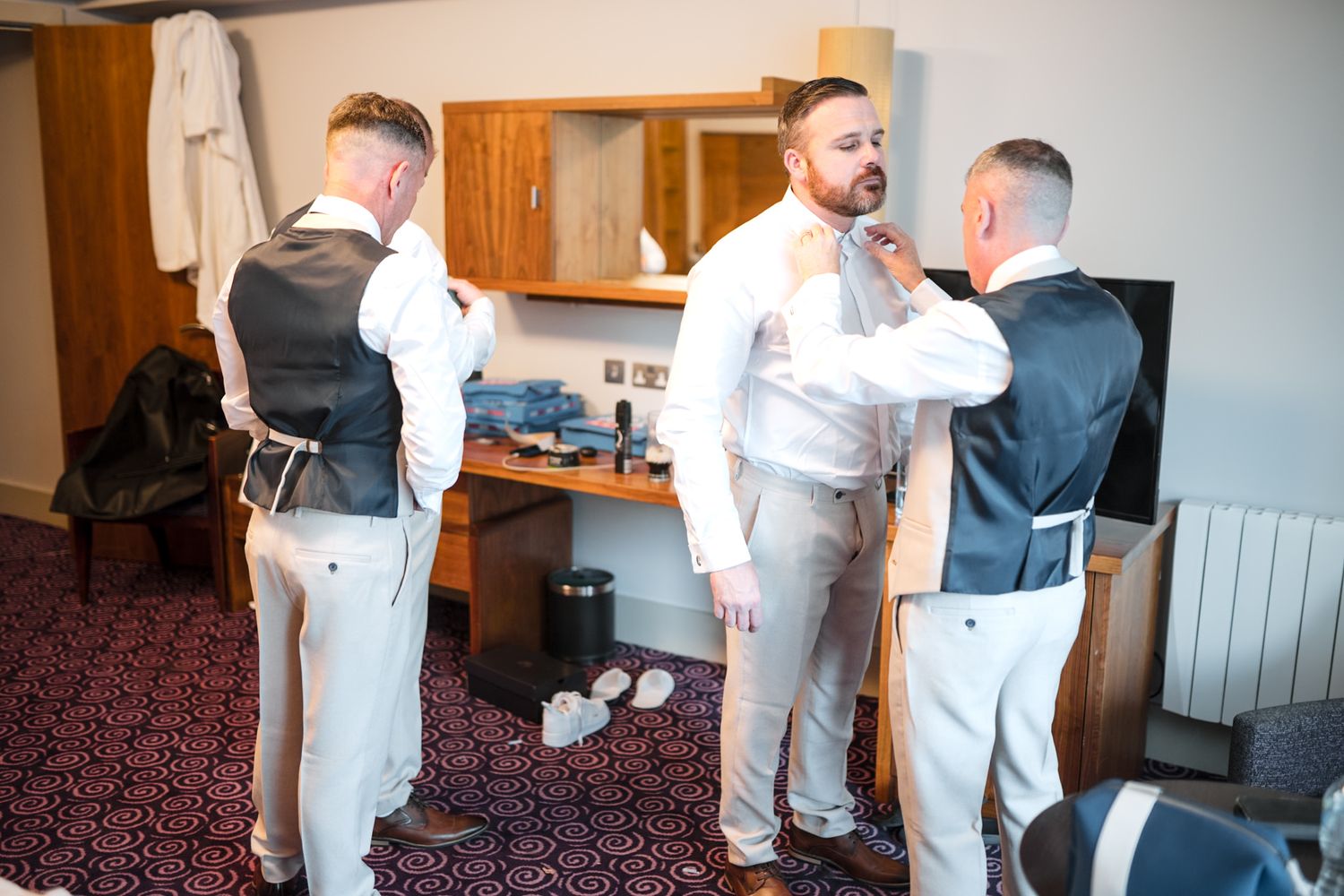 Groomsmen getting dressed in white shirts and formal attire in a hotel room before a wedding ceremony.