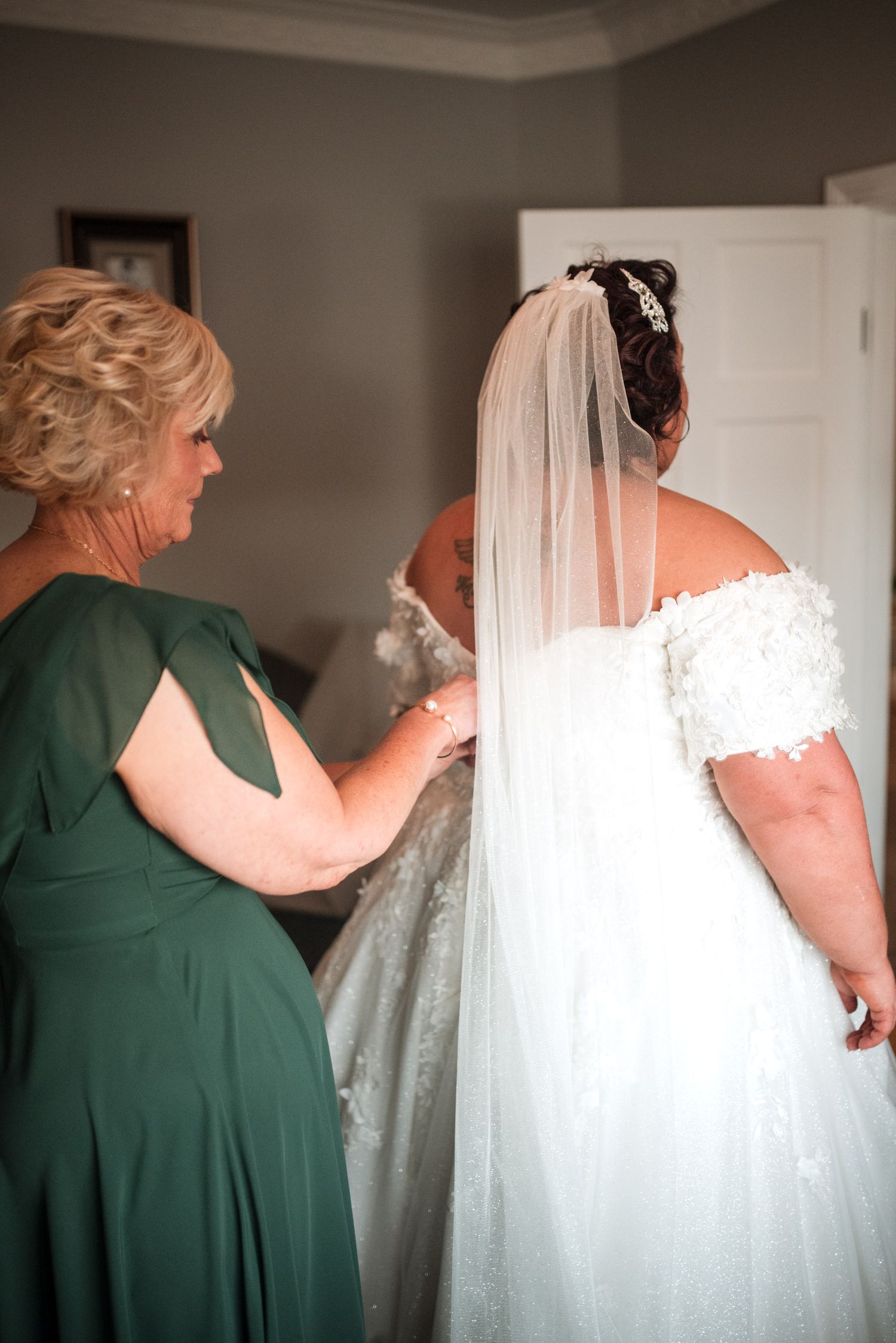 A person in a green dress helps adjust a bride's lace wedding gown and long white veil during wedding preparations.
