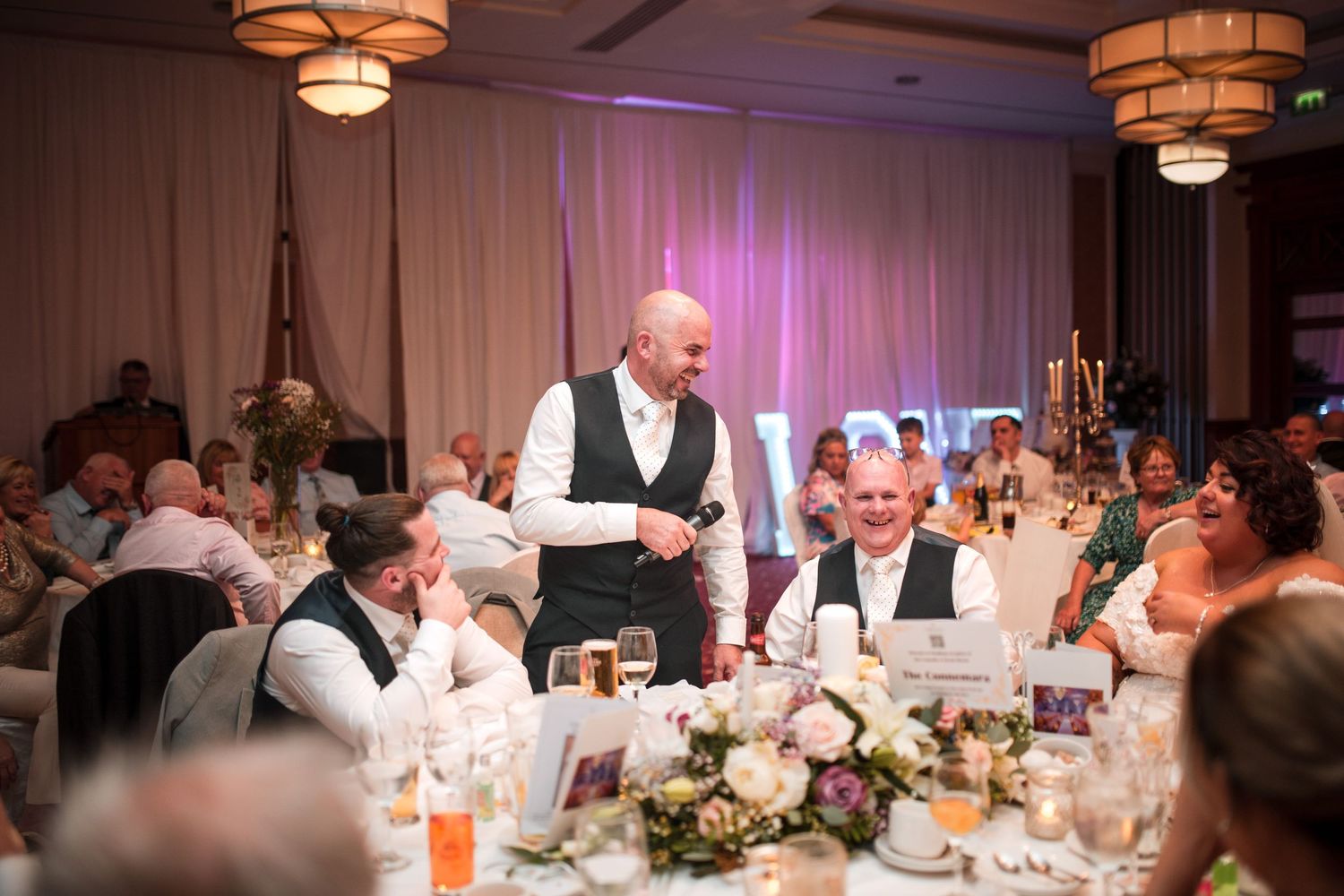 A waiter in a white shirt and black vest serves guests at an elegant wedding reception with soft purple lighting and floral centerpieces.