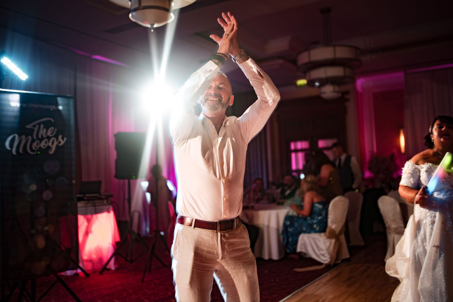 Person in light-colored attire dances enthusiastically with raised arms on a dance floor with pink and purple lighting.