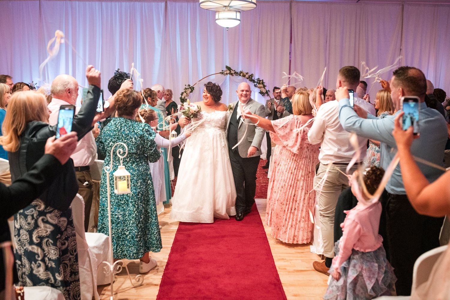 Wedding guests raise their arms in celebration as newlyweds walk down red carpet aisle in elegant ballroom reception.