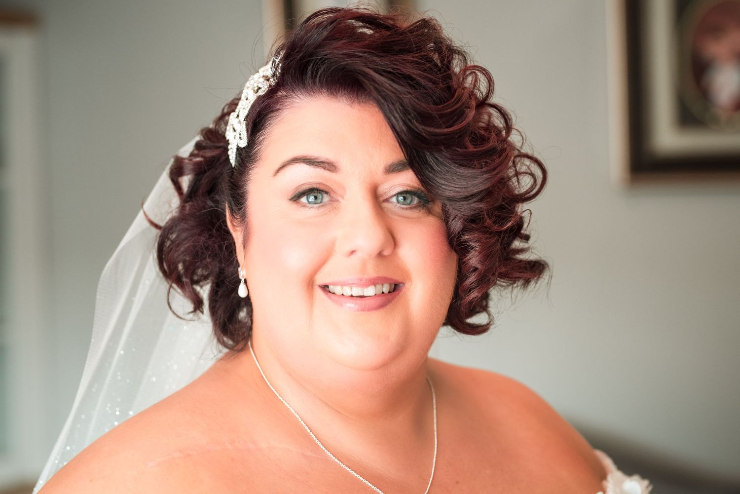 A bride in a shoulder strap dress and silver necklace smiles warmly while wearing her hair in dark curls.
