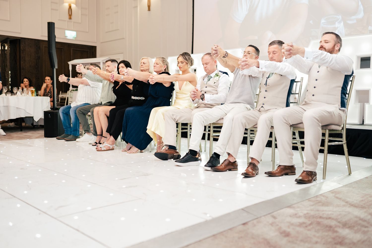 People sitting in a row against a white wall perform synchronized arm movements during a group activity.