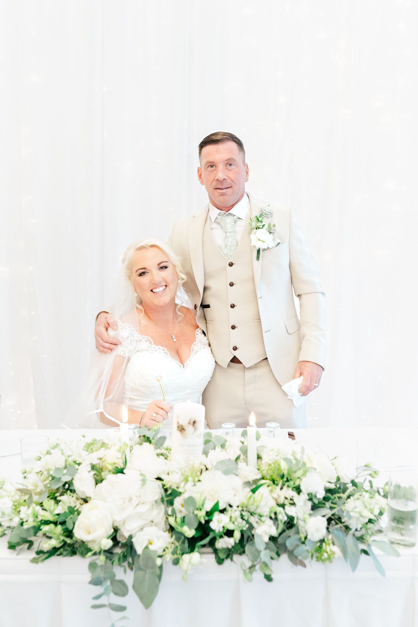 Newlyweds share intimate moments at head table decorated with lush white floral arrangements and greenery.
