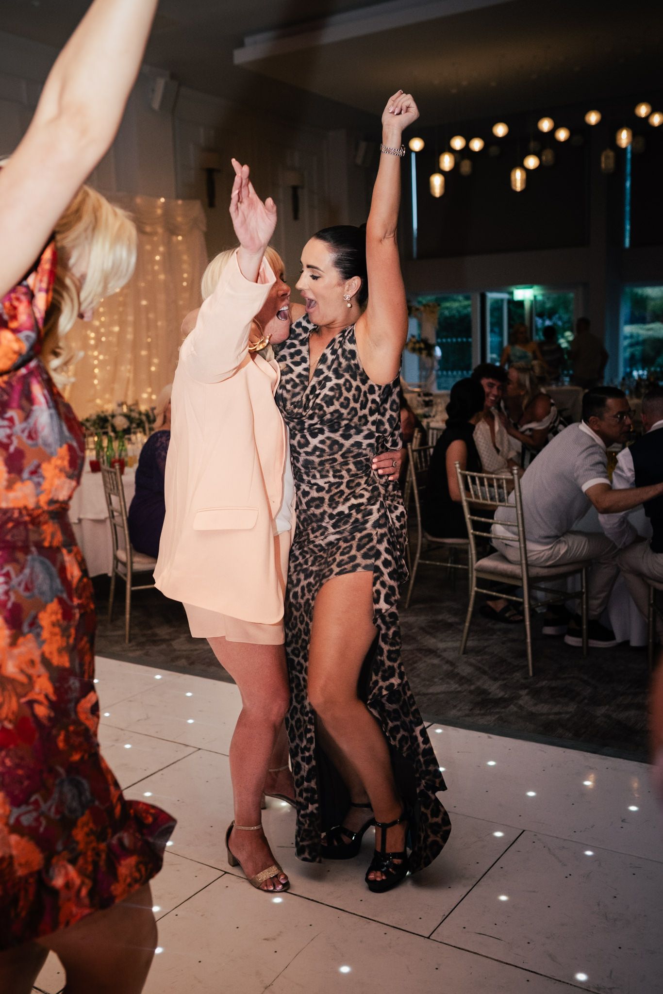 Partygoers dancing enthusiastically at an indoor wedding reception with string lights and decorations visible in background.