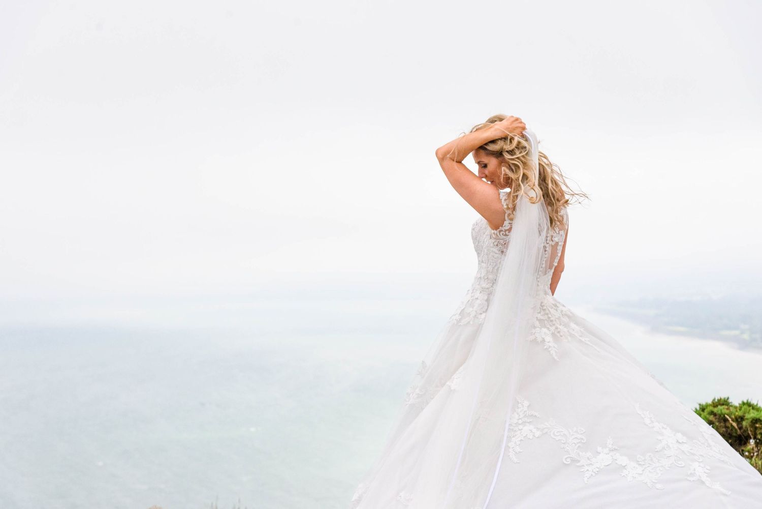 A person in a flowing white wedding dress twirls on a cliff overlooking a misty ocean view.