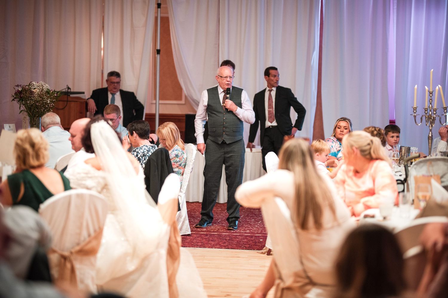 Wedding reception guests dancing to music on a dance floor with reception tables and ballroom decor visible.