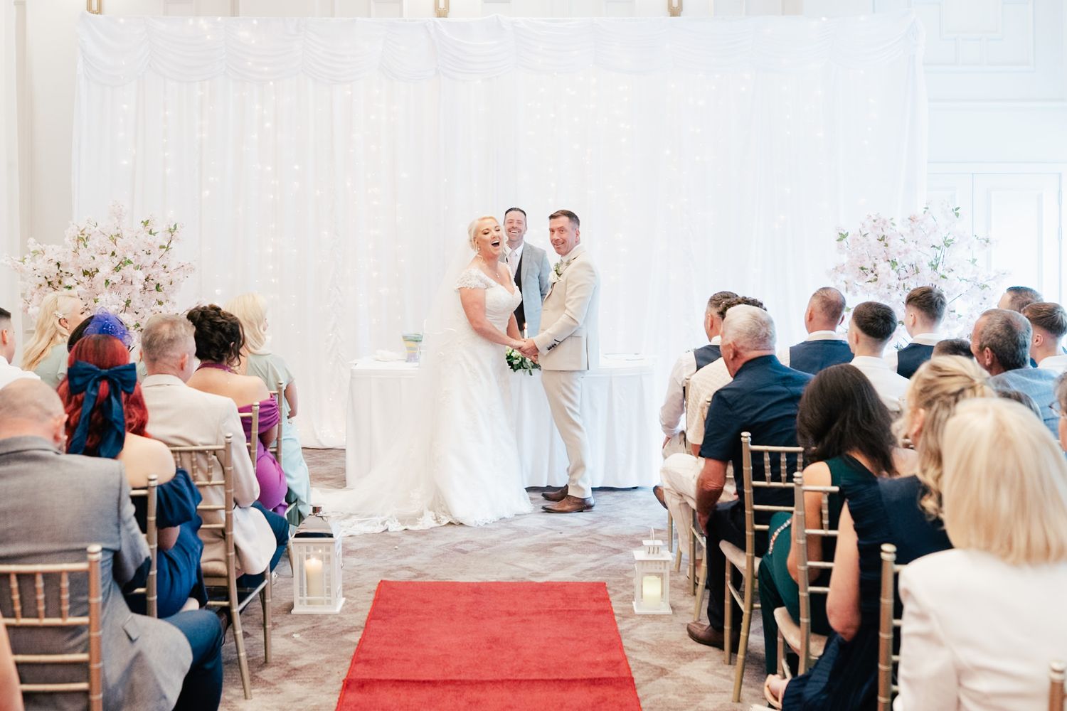 Wedding ceremony in a bright white room with a red carpet aisle and guests seated in chiavari chairs.