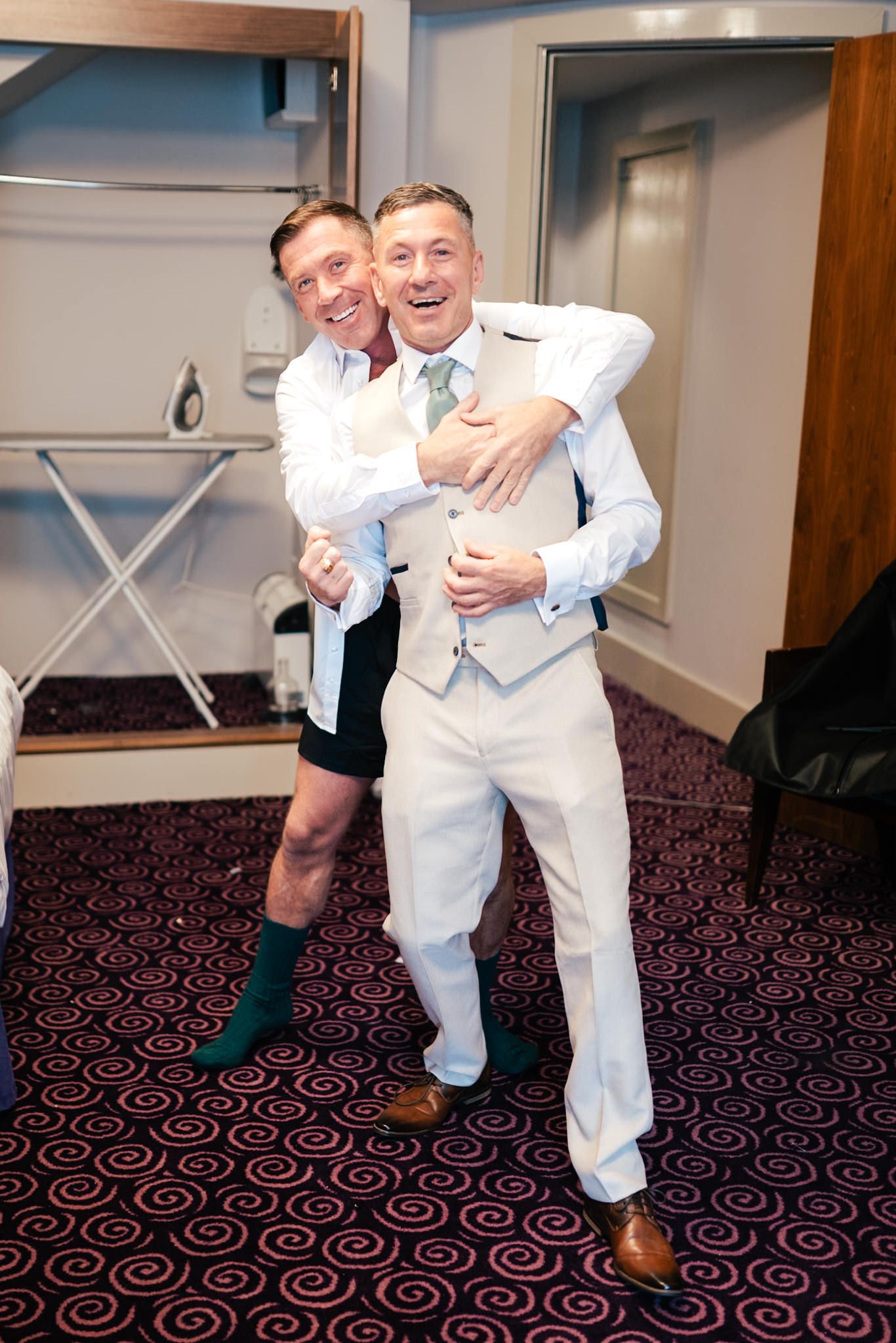 Two people share a joyful embrace while posing in a room with patterned carpet and white walls.