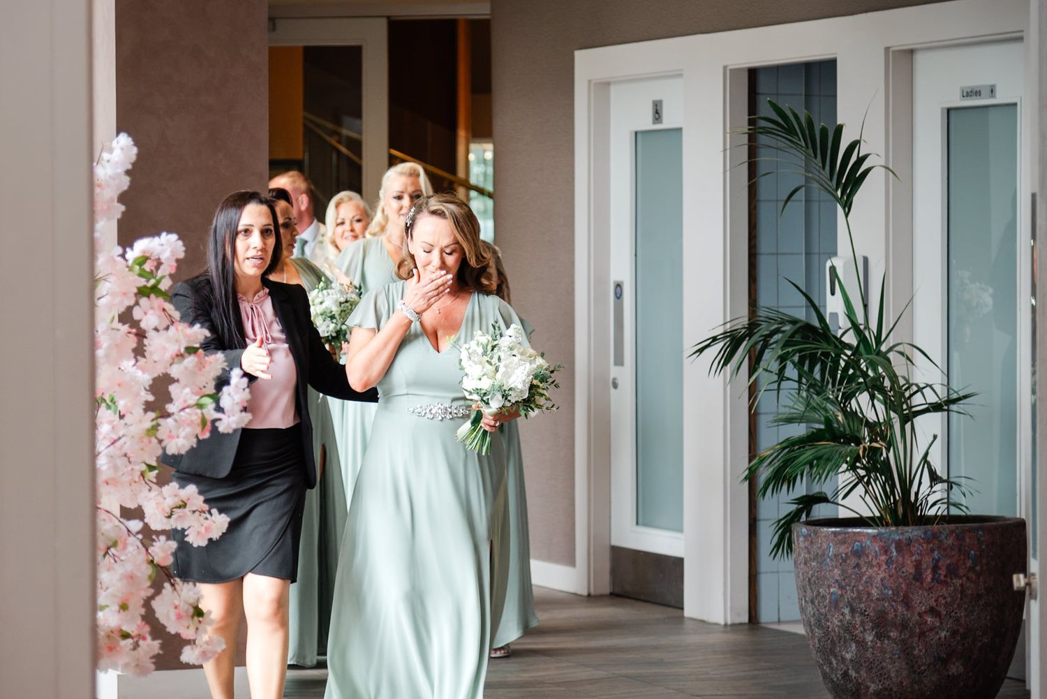 A group poses elegantly in formal attire near a mint green wall and decorative cherry blossom arrangement.