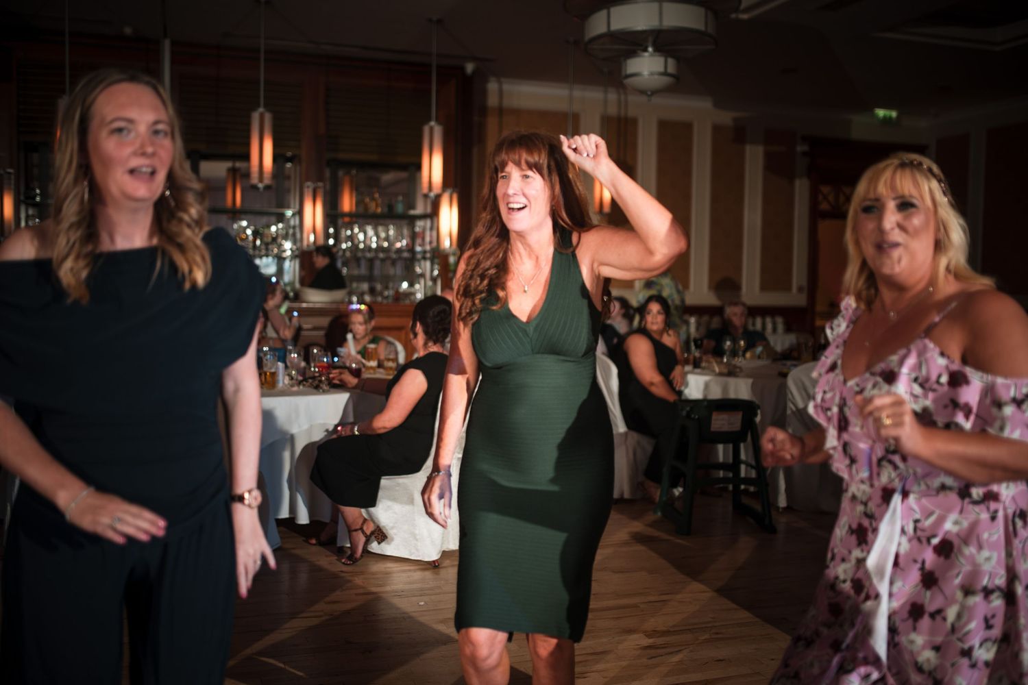 Three people happily dancing together at an indoor reception with tables and lighting in the background.