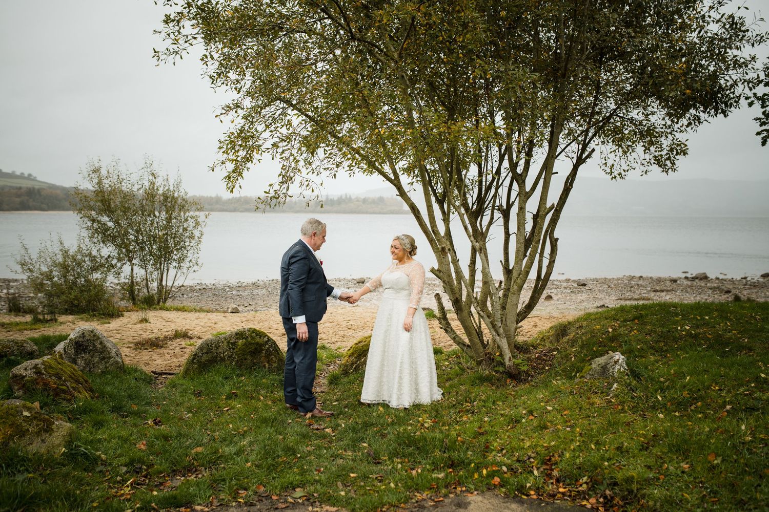 A couple shares a tender moment on a scenic coastal shoreline beneath a tree on an overcast day.