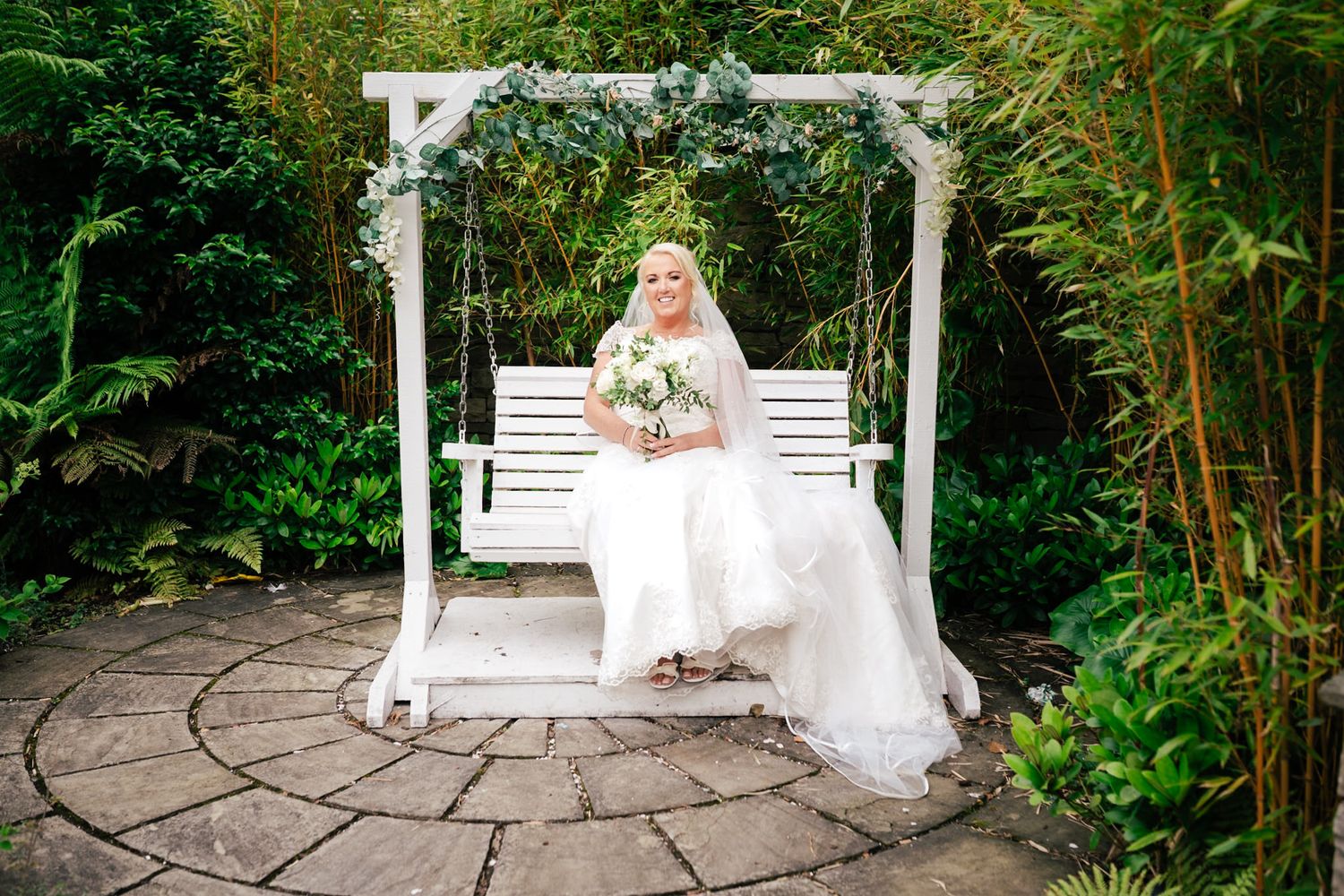Elegant bride poses gracefully on a white garden bench surrounded by lush greenery and natural garden landscaping.
