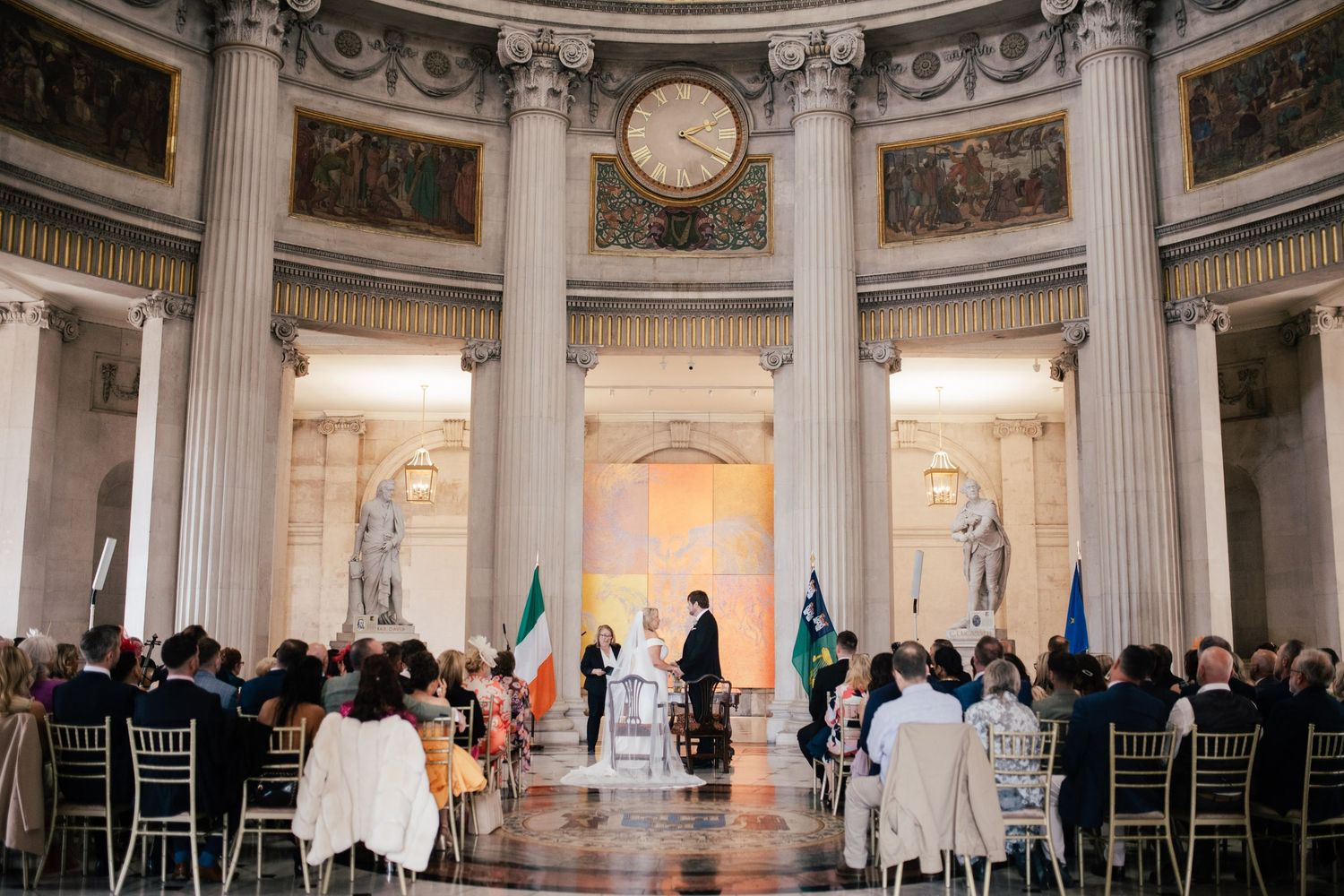 Elegant wedding ceremony taking place in a grand neoclassical rotunda with marble columns and seated guests.