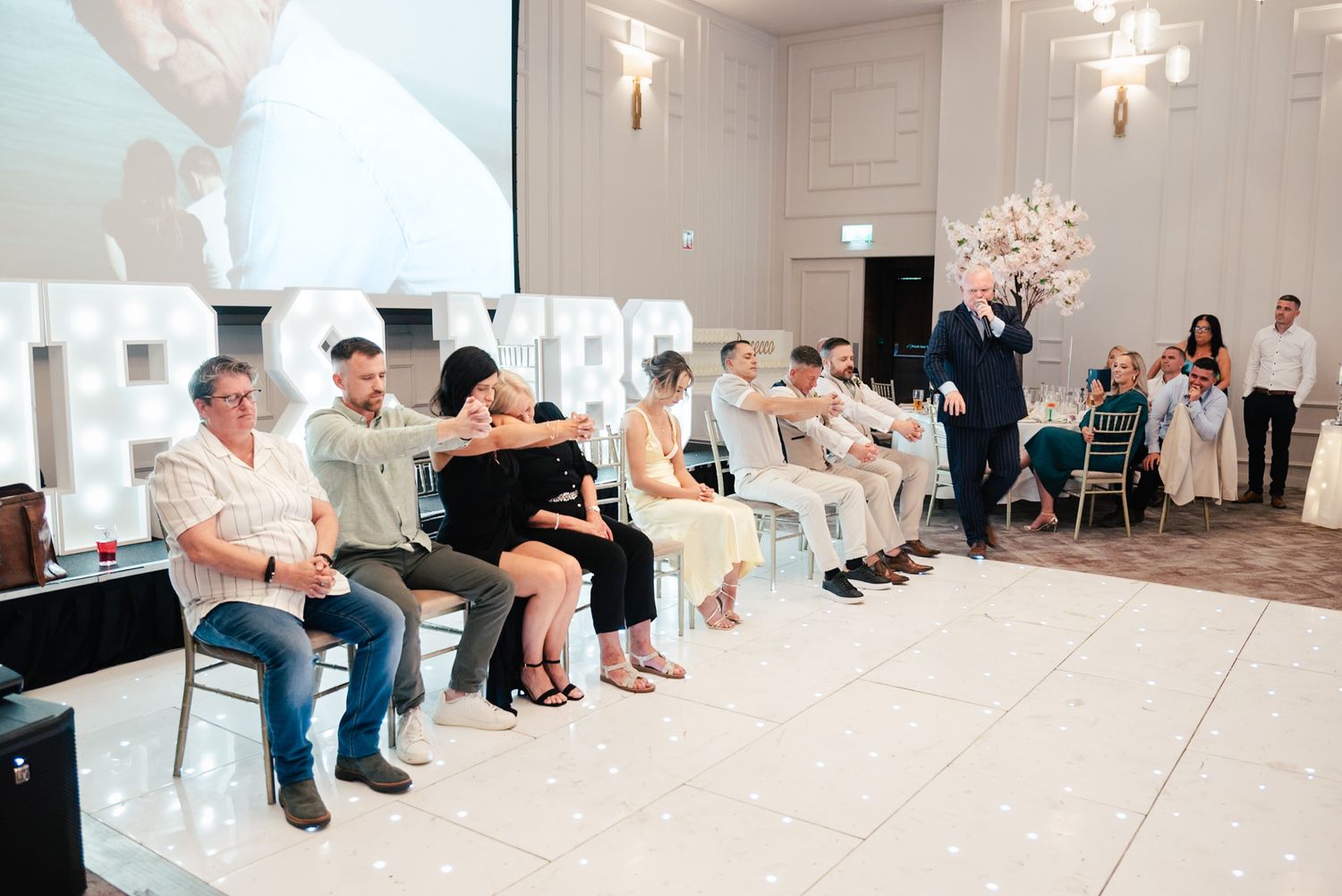 Group of party guests sitting in a row near large decorative letters at a light-colored indoor venue.